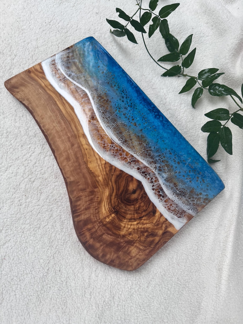 Pode incluir: Uma t&aacute;bua de servir de madeira com um design de resina ep&oacute;xi azul e branco que se assemelha a ondas do oceano. A t&aacute;bua tem uma veia de madeira natural e uma borda curva.