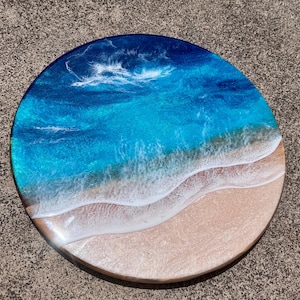 May include: Circular resin artwork showing an ocean scene. The piece uses layers of blue and turquoise resin to represent water, with white resin waves breaking on a sandy beach. This is a decorative item.