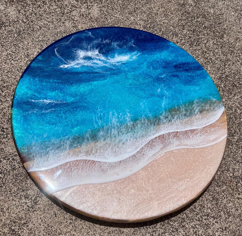 May include: Circular resin artwork showing an ocean scene. The piece uses layers of blue and turquoise resin to represent water, with white resin waves breaking on a sandy beach. This is a decorative item.