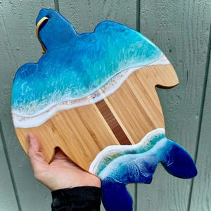 May include: A blue and green resin turtle-shaped cutting board with a wood center. The resin is designed to look like ocean waves.