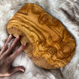 Pode incluir: Uma t&aacute;bua de cortar de madeira de oliveira grande, de forma oval, com um padr&atilde;o de gr&atilde;o de madeira natural. A t&aacute;bua tem uma cor castanho claro com veios e n&oacute;s castanhos mais escuros.