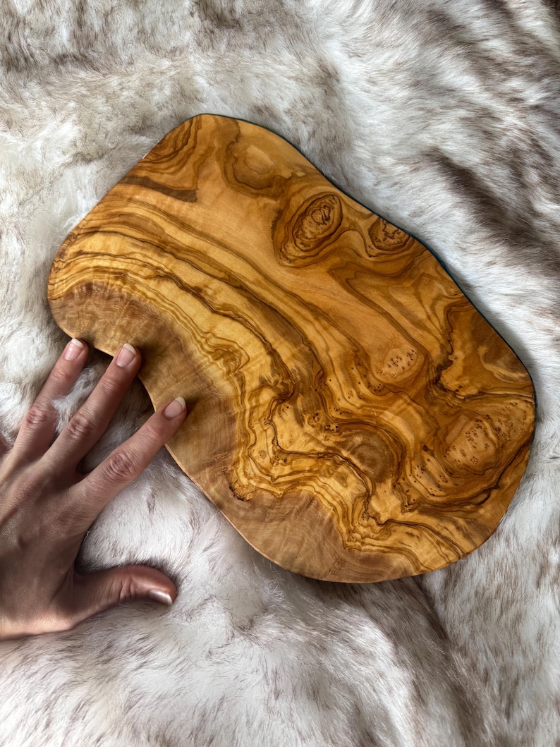 Pode incluir: Uma t&aacute;bua de cortar de madeira de oliveira grande, de forma oval, com um padr&atilde;o de gr&atilde;o de madeira natural. A t&aacute;bua tem uma cor castanho claro com veios e n&oacute;s castanhos mais escuros.