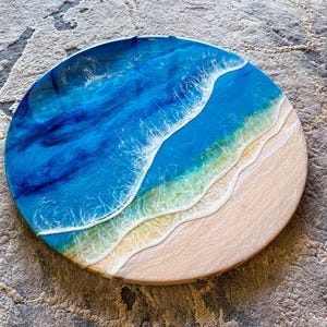 May include: Circular resin artwork showing an ocean scene. The piece uses layers of blue and white resin to create the effect of waves. The bottom section is a sandy beach colour. The diameter of the piece is approximately 30 cm.