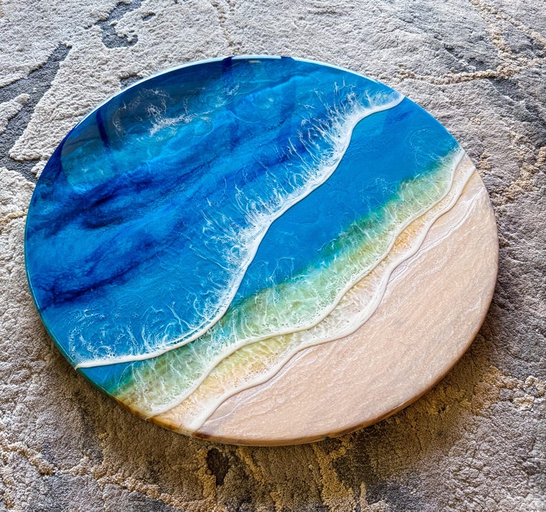 May include: Circular resin artwork showing an ocean scene. The piece uses layers of blue and white resin to create the effect of waves. The bottom section is a sandy beach colour. The diameter of the piece is approximately 30 cm.