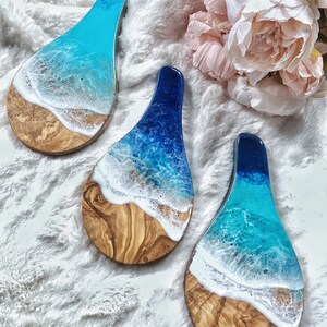 May include: Three wooden serving boards with a resin ocean design. The boards feature a light brown wood base with a blue and white resin wave design. The resin colors range from light blue to deep blue, with white foam detailing. The boards are teardrop shaped.