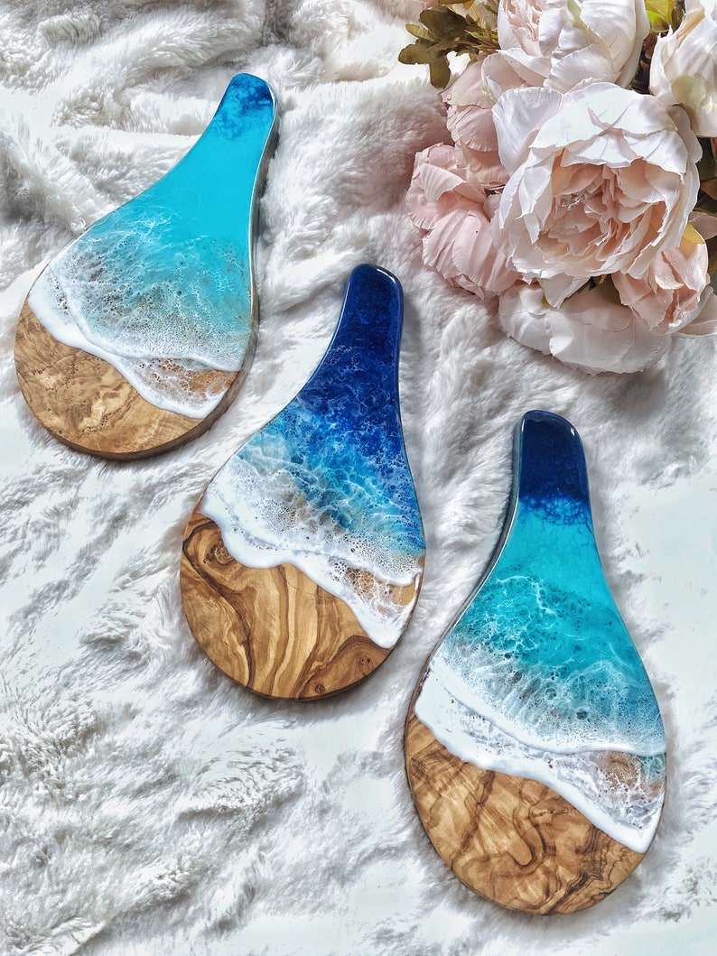 May include: Three wooden serving boards with a resin ocean design. The boards feature a light brown wood base with a blue and white resin wave design. The resin colors range from light blue to deep blue, with white foam detailing. The boards are teardrop shaped.