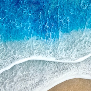 May include: Abstract artwork of a beach scene. The image shows a gradient of colours, from deep blue to light blue, representing the ocean. White foamy waves meet a tan sandy shore. The artwork evokes a sense of tranquility and the beauty of nature.