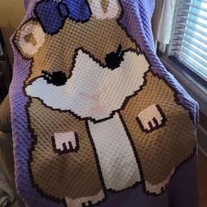 May include: A crocheted blanket featuring a brown and white hamster with a purple bow. The blanket is 4.5 feet by 6 feet and was made with a 4.00 mm hook. The text "Picture courtesy of Andi Rogers" is at the bottom of the image.