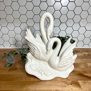 May include: A white ceramic planter shaped like two swans with their necks curved toward each other. The base features a molded water lily design. A green plant with patterned leaves is tucked into the center of the swan sculpture.