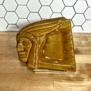 May include: A glazed ceramic wall pocket or plaque in a mustard yellow color featuring a molded relief of a person wearing a feathered headdress. The piece has a glossy finish and is supported by a thin, curved gold-toned metal wire stand.