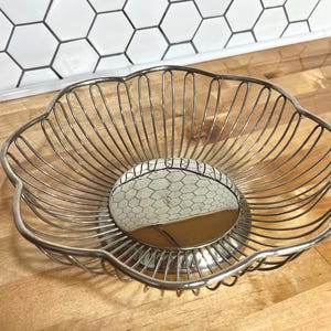 May include: A silver-colored metal wire basket with a flower-shaped rim. The basket features a solid, reflective circular base and thin, curved wire sides that extend upward to form a scalloped edge, creating an open, airy structure for storage.
