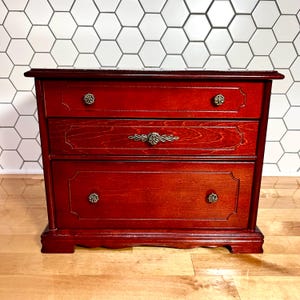 May include: A small, dark reddish-brown wooden chest with three horizontal drawers. The top and bottom drawers feature two round, ornate metal knobs each, while the middle drawer has a single elongated, decorative metal handle in the center.