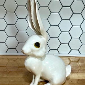 May include: A white ceramic rabbit figurine with a glossy finish, featuring long upright ears and large, dark amber-colored eyes. The rabbit is depicted in a seated position with its front paws resting on the ground and a small tail visible.