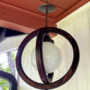 May include: A hanging pendant light fixture featuring a spherical white glass globe encased within two intersecting dark brown wooden rings. The fixture is suspended by a metal chain and a circular ceiling canopy, with a small finial at the bottom.