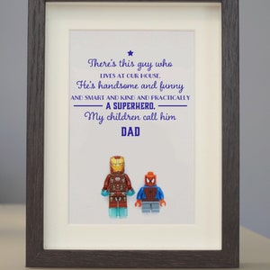 May include: A framed print with a white background and a blue text message that reads "There's this guy who lives at our house. He's handsome and funny and smart and kind and practically a superhero. My children call him DAD".  Two small toy figures are at the bottom of the print, one is red and blue and the other is red and gold.