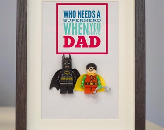 Puede incluir: Una impresión enmarcada con el texto "Quién necesita un superhéroe cuando tienes a papá" en letras rojas, azules y turquesa. Dos minifiguras de Lego, una vestida de Batman y la otra de Robin, están unidas a la impresión.