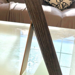 May include: A dark brown wooden picture frame with a stand on the back. The frame is standing on a glass table.
