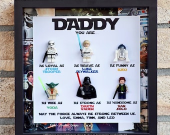May include: A framed picture with a white background and a collage of Star Wars characters. The text reads "Daddy You Are..." and lists qualities like "Loyal as Stormtrooper", "Brave as Luke Skywalker", "Funny as R2D2", "Wise as Yoda", "Strong as Darth Vader", and "Handsome as Han Solo". The text also includes "May the Force Always Be Strong Between Us. Love, Emma, Finn, and Leo".