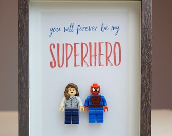 May include: A framed print with the text "you will forever be my SUPERHERO" in red and blue lettering. Two Lego minifigures are placed below the text, a female minifigure with brown hair and a male minifigure dressed as Spiderman.