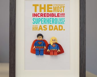 May include: Framed print with a colorful word art design that reads "The strongest, most incredible and daring superhero lives here. We know him as Dad."  Two Lego minifigures of Superman and Superwoman are displayed in the frame.