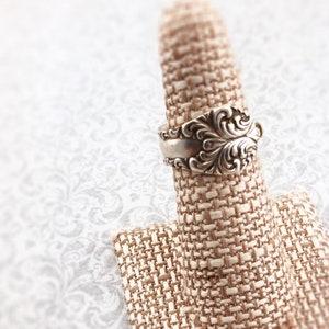 May include: A silver spoon ring with an ornate design. The ring is displayed on a beige fabric ring holder.