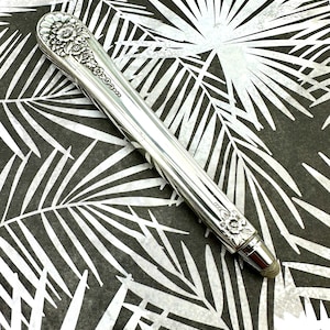 May include: A silver stylus with a floral design on the top and bottom. The stylus is on a black and white background.