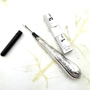 Puede incluir: Un enhebrador de aguja de plata con un mango adornado y una funda de plástico negro. El enhebrador mide aproximadamente 12,5 cm de largo y está al lado de una cinta métrica de la marca Singer.