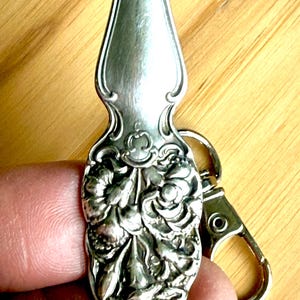 May include: Silver-toned spoon keychain with ornate floral detailing. The spoon's handle features a decorative floral design, and it is attached to a silver-colored clasp. The keychain is held by a person.