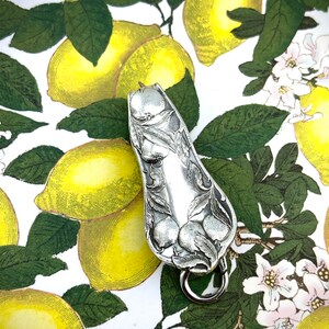 Könnte beinhalten: Ein silberner, löffelförmiger Schlüsselanhänger mit einem verzierten Design aus Früchten und Blättern. Der Schlüsselanhänger hat einen kleinen Metallring am unteren Ende. Der Hintergrund zeigt ein Muster aus Zitronen und grünen Blättern.