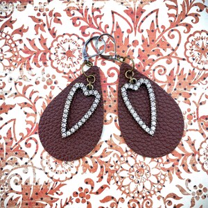 Può includere: Un paio di orecchini in pelle marrone a forma di lacrima con un design a cuore di strass.