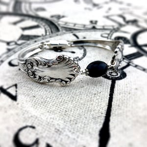May include: A silver spoon bracelet with a black bead charm. The bracelet is on a fabric background with a clock face pattern.