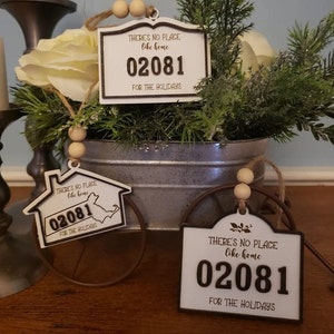 May include: Three wooden ornaments with the text "There's no place like home 02081 for the holidays". The ornaments are white with brown text and are shaped like a house, a sign, and a sign with a rounded top.