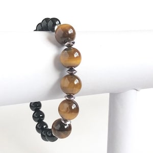 May include: A beaded bracelet with alternating black and tiger's eye beads. The bracelet is displayed on a white stand.