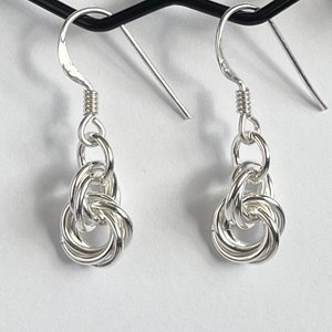 May include: A pair of silver earrings with a knot design. The earrings are hanging from a black stand.