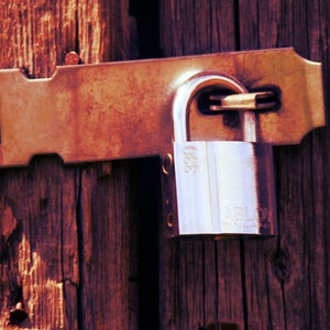 Puede incluir: Un candado plateado con el texto "ABLOY FINLAND" y "035" en él está unido a un pestillo de metal oxidado en una puerta de madera.