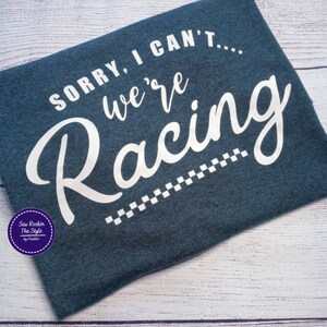 May include: A dark blue t-shirt with the text "SORRY, I CAN'T... we're Racing" in white script. A checkered flag design is below the word "Racing". The shirt also has a circular logo in the lower left corner.