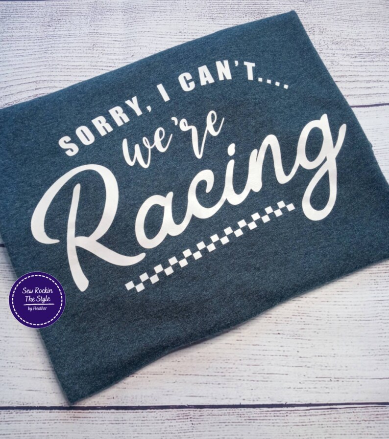 May include: A dark blue t-shirt with the text "SORRY, I CAN'T... we're Racing" in white script. A checkered flag design is below the word "Racing". The shirt also has a circular logo in the lower left corner.