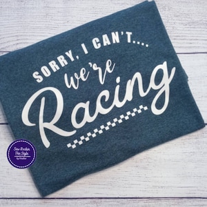 May include: A navy blue t-shirt with the text "SORRY, I CAN'T... we're Racing" in white script. A checkered flag design is at the bottom. The shirt has a crew neck and is made of a soft material.