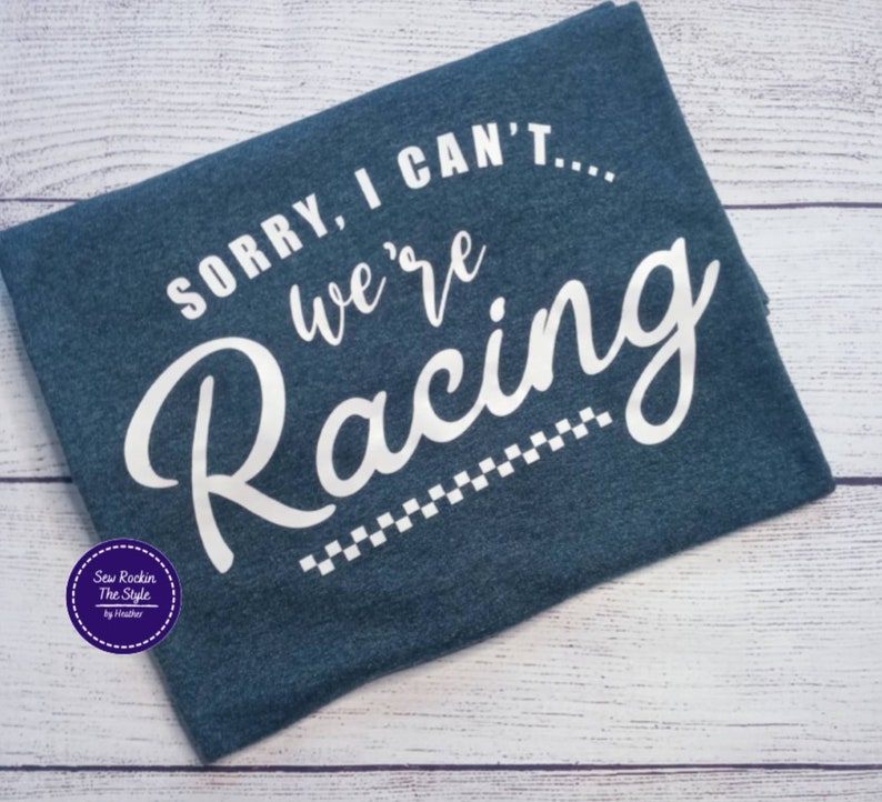 May include: A navy blue t-shirt with the text "SORRY, I CAN'T... we're Racing" in white script. A checkered flag design is at the bottom. The shirt has a crew neck and is made of a soft material.