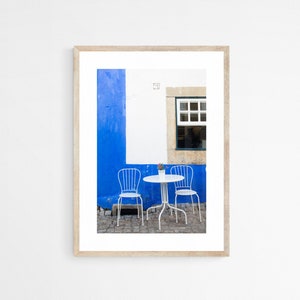 A pair of white iron cafe chairs at a little white table against a bright blue wall with an open window in Obidos, Portugal