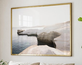 May include: A light beige sectional sofa with two cushions sits in a living room with a large window, a wooden coffee table, a woven chair, and a potted plant with green leaves. A framed photograph of a seascape hangs on the wall above the sofa.