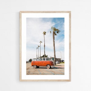 A vertical orientation photograph of an orange vintage Volkswagen bus in a parking lot with palm trees behind it and a seagull flying in the sky.