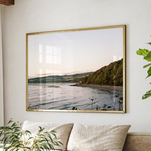 May include: A modern living room with a beige sectional sofa, a wooden coffee table, and a woven chair. A large framed landscape art piece hangs above the sofa. A large green plant sits in a woven basket. White curtains and a window are visible.