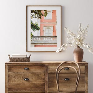 A bedroom scene looking straight on at a wood dresser decorated with a brown vase and a basket. over the dresser hangs a wood frame with a white mat and a photograph of a coral pink building with a white stone balcony and white windows and leaves from a magnolia tree framing the left side of the composition. The wall behind is white.