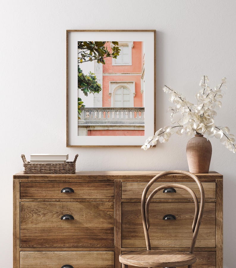 A bedroom scene looking straight on at a wood dresser decorated with a brown vase and a basket. over the dresser hangs a wood frame with a white mat and a photograph of a coral pink building with a white stone balcony and white windows and leaves from a magnolia tree framing the left side of the composition. The wall behind is white.