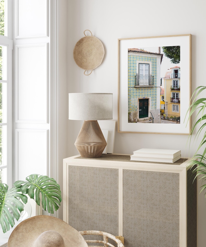 May include: A framed print of a building with blue and white tile work in Lisbon, Portugal. The building has a green door and a balcony with a wrought iron railing. The print is hanging on a white wall above a wooden cabinet with a woven rattan front.