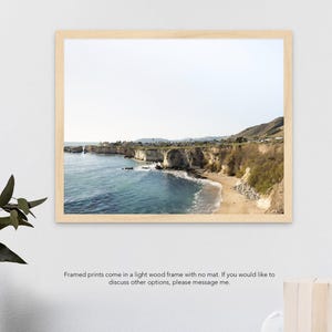 May include: A framed photograph of a rocky coastline with a sandy beach and blue ocean water. The photo is in a light wood frame with no mat.