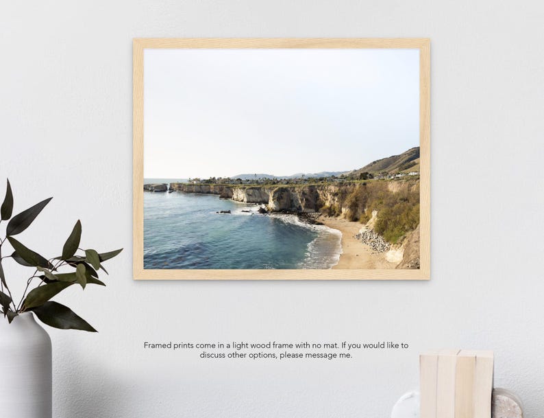 May include: A framed photograph of a rocky coastline with a sandy beach and blue ocean water. The photo is in a light wood frame with no mat.