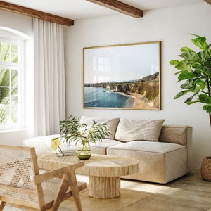 May include: A light beige sectional sofa with a large framed photograph of a coastal scene above it. A potted ficus tree is on the right side of the sofa. A wooden coffee table with a woven top sits in front of the sofa. A woven chair is in the foreground.