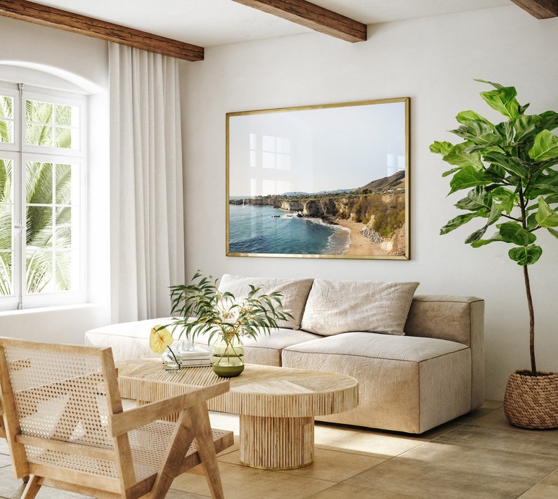 May include: A light beige sectional sofa with a large framed photograph of a coastal scene above it. A potted ficus tree is on the right side of the sofa. A wooden coffee table with a woven top sits in front of the sofa. A woven chair is in the foreground.